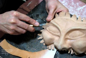 leather unfinished mask, hand holding a small sharp knife cutting the right eye out of his mask