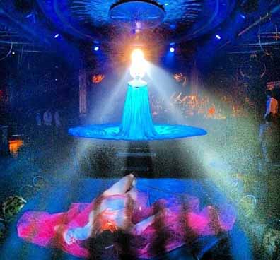 Queen of the Night Mask,  photo of Katherine Crockett standing in a blue dress with a lazer light hitting the stone at the top of the mask and reflecting throughout the theater.