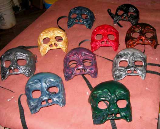 10 finished skull masks, green, gray silver, light blue, silver, yellow, blue, red , black and browish red all laying on a table in my workshop
